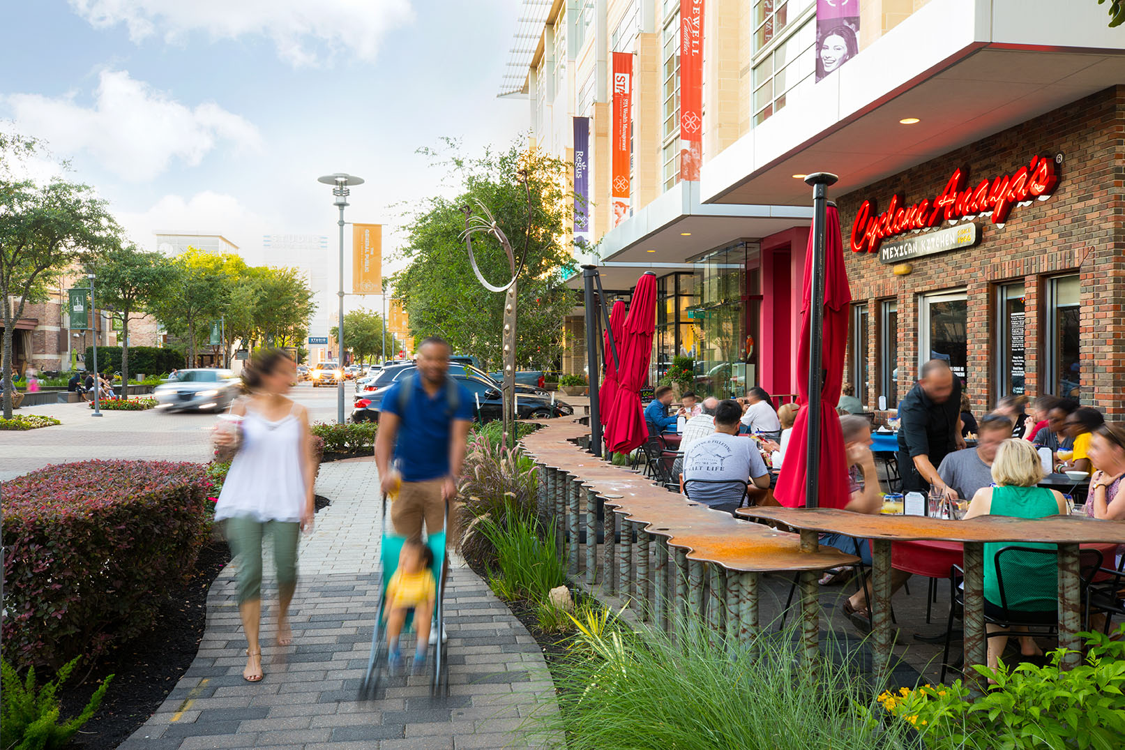 CITYCENTRE promenade at golden hour, with families walking past patio dining, planters, and tree-lined pavers on Houston's west side